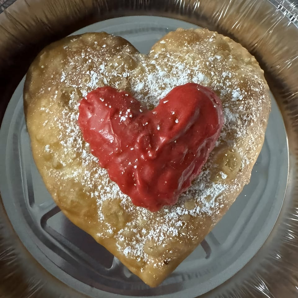 Heart-shaped fried dough with red icing and powdered sugar in a foil container.