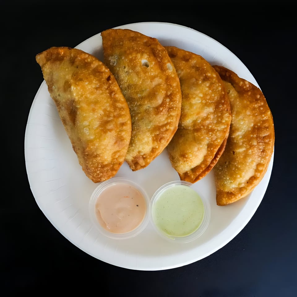 Four golden-brown empanadas and two dipping sauces on a white plate against a dark background.